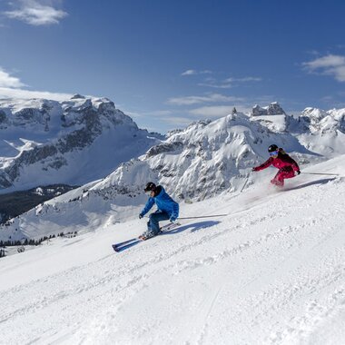 Zwei sportliche Skifahrende fahren eine verschneite Piste hinab, umgeben von schneebedeckten Bergen. | © Stefan Kothner - Montafon Tourismus GmbH