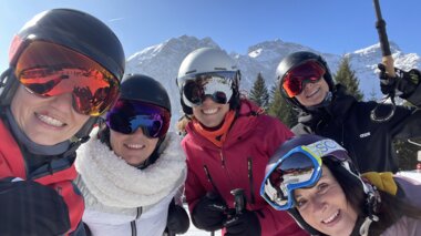 Fünf Damen machen ein gemeinsames Gruppenbild auf der Piste während eines Skitages, im Hintergrund sind die Berge und der blaue Himmel zu sehen. | © Melanie Fleisch