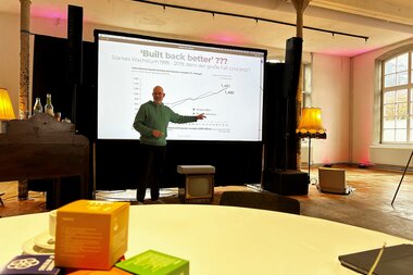 Ein Vortragender bei einer nachhaltigen Veranstaltung erklärt die Grafik auf einer Folie, die hinter ihm projeziert ist. | © Anna Engstler