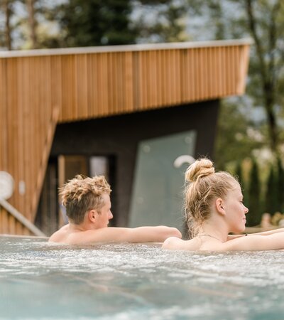 Zwei Personen, ein Mann und eine Frau, entspannen im Outdoor-Pool des Wellnessbereichs im Val Blu; im Hintergrund Garten, Bäume und eine Sauna bei Tageslicht. | © Hefti Fotografie