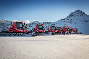 Acht PistenBullys stehen im Skigebiet Brandnertal in einer Reihe. Im Hintergrund, ein Bergpanorama. | © Bergbahnen Brandnertal, Michael Marte