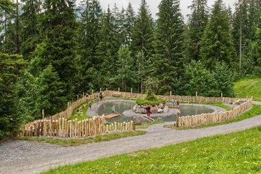 Spielplatz am See mit Floß, Balancier-Baumstamm und weiteren Spielstationen, eingebettet in die Natur; vorne eine Wiese, dahinter ein Weg und Bäume im Hintergrund. | © Seilbahnen Sonntag-Stein